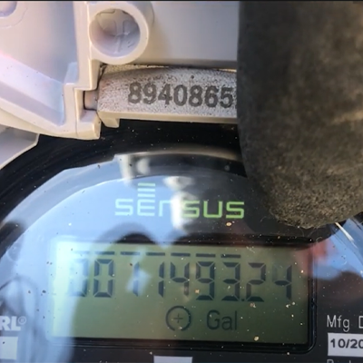 Close-up image of AMI water meter with gloved finger pointing at the meter's flow indicator.